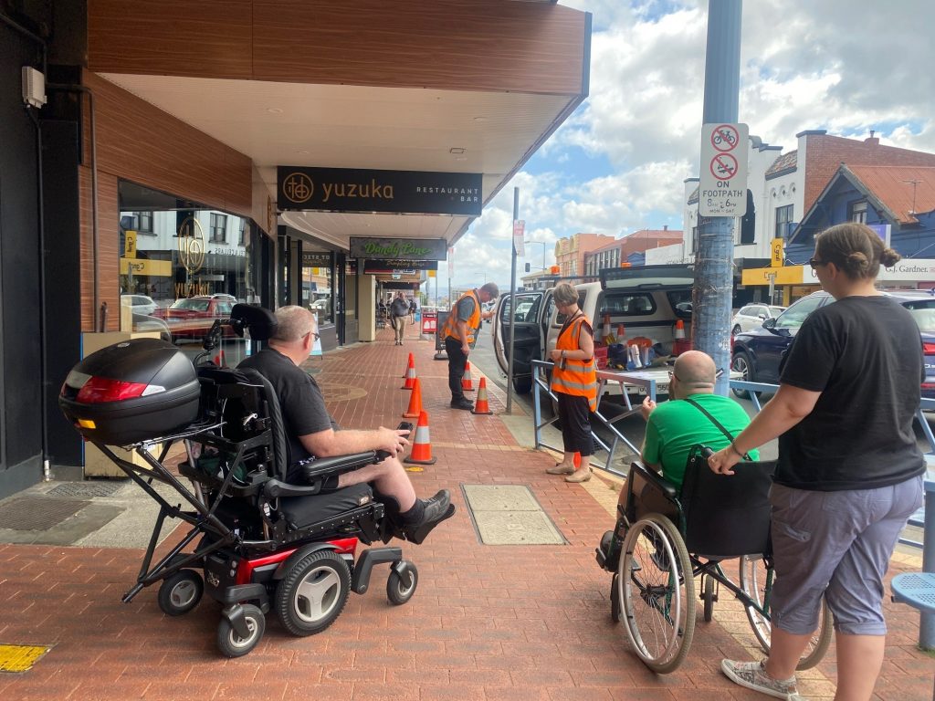 Council officers mark out the trading zone with orange cones. Members of the Access group are watching. THe Council officer is holding a vehicle door open next to the kerb.